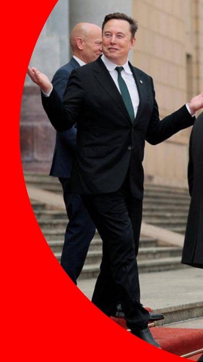 Elon Musk stands on a red carpet and wears a suit.