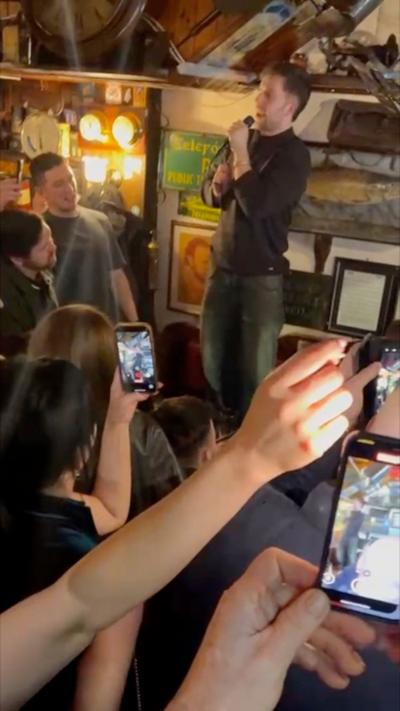 A man holding a microphone, stood on a pub table while a large crowd films him