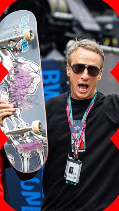 Tony Hawk holding up a skateboard, wearing black sunglasses.