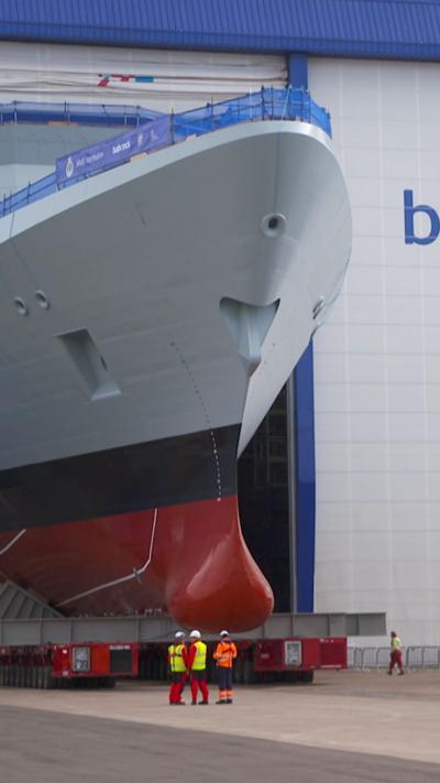 The HMS Venturer emerges from a blue and white hall with Babcock written on the side. It is a grey, black and red warship. Bystanders wearing high vis jackets and helmets look on.