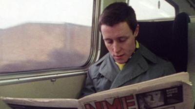 A man reads an NME newspaper on a train.
