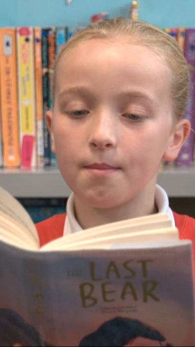 A young girl with blond hair tied back in a ponytail is reading a book called The Last Bear. We can partially see the front cover, which depicts a bear and a young girl. The reader is sitting in front of a bookshelf lined with books sitting in front of a blue wall. She is wearing a red school uniform jumper over a white polo shirt. 