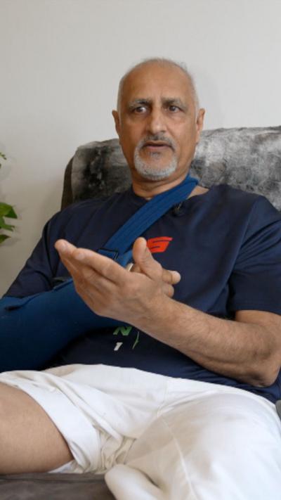 A balding man with a blue t-shirt and white shorts. His right arm is in a sling