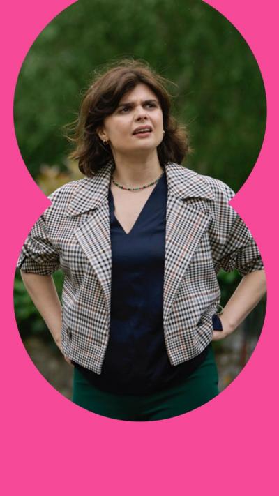 Character DS Janie Mallowan stands outdoors looking puzzled; she wears a checked jacket and has medium length dark hair