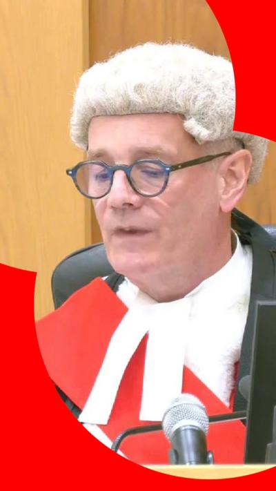 A judge seen wearing a wig, robes and glasses, reads from his laptop