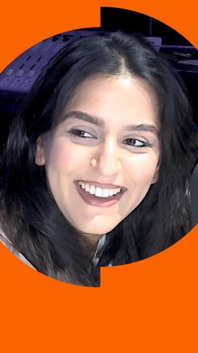 A woman with long black hair sits in front of a microphone. Her head is turned to look off camera and she is smiling.