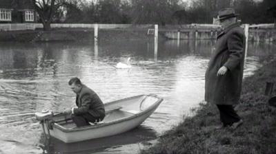 A man in a fibreglass boat, watched by a man on the riverbank.