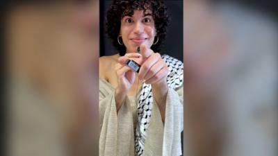 Jacon Alon points at the camera, wearing a flowy silver top. They have curly black hair and silver earrings 