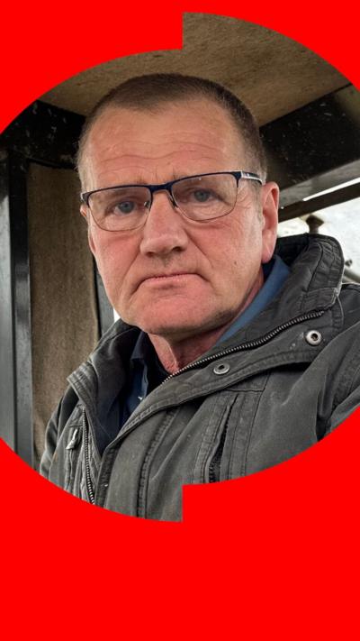 A man with short black hair sits in a tractor cab. He is wearing glasses and heavy-duty dark grey coat.
