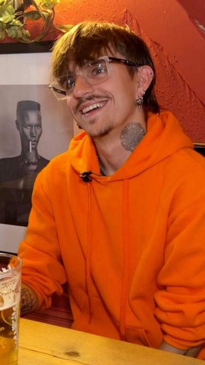 A man with brown hair and glasses, and a neck tattoo sat in a pub. He is wearing an orange hoodie