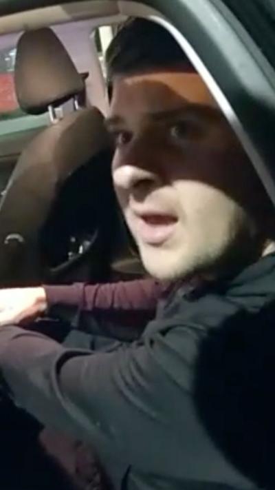 A man sat in the back sear of a black car is looking up at the camera, there is a shadow across his face and his mouth is open. He is wearing a black jacket.