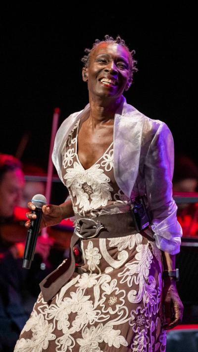 Live concert performance featuring a female vocalist in an embroidered gown smiling on stage with microphone and orchestra behind