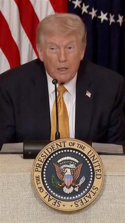 Donald Trump sits down in front of a US flag and speaks into a microphone with a US Presidential Seal emblem in front of him.