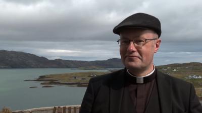 Maighstir Ross na sheasamh le ad air agus gàire air aodann. 