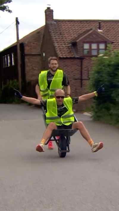 Two men are competing in a wheelbarrow race, both in yellow hi-vis jackets. One man pushes another man who sits in the wheelbarrow with his arms outstretched wide, and wears black sunglasses and gloves. He is bald. The man pushing has brown hair, and his tongue out towards the camera. Behind the men is a residential area, with brown brick houses, green shrubbery, wheelie bins and orange cones surrounding them. The floor beneath them is a light grey concrete.