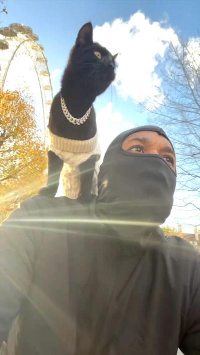 A man wearing a balaclava rides a bike, passing to the London Eye. He has a black cat on his shoulder, which is wearing a silver chain around it's neck..