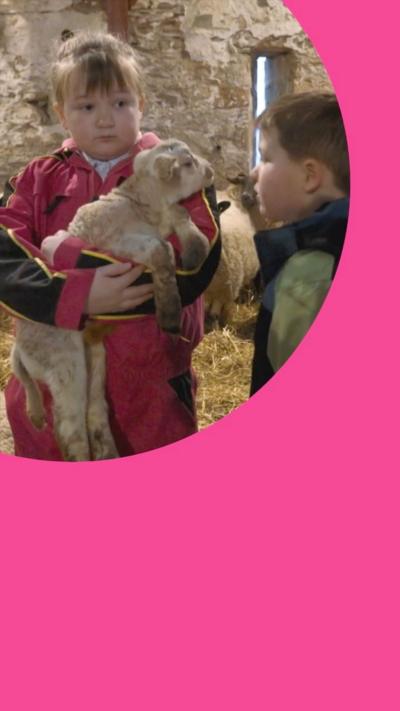 Two children in barn, one holding a baby lamb.