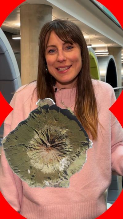 A woman in a pink jumper stands in an office. A top-down view of a volcano is superimposed in front of her