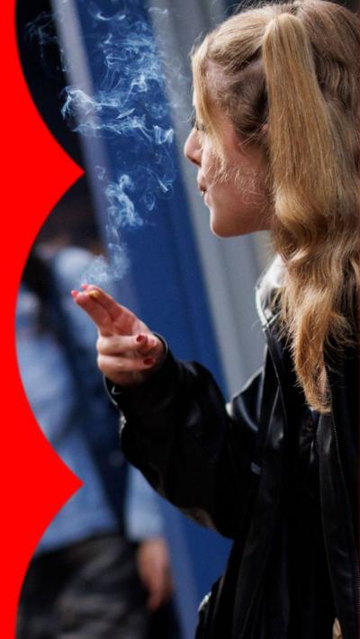 A woman with long, wavy blonde is standing outside smoking a cigarette. Behind her, another woman walks out through a doorway.