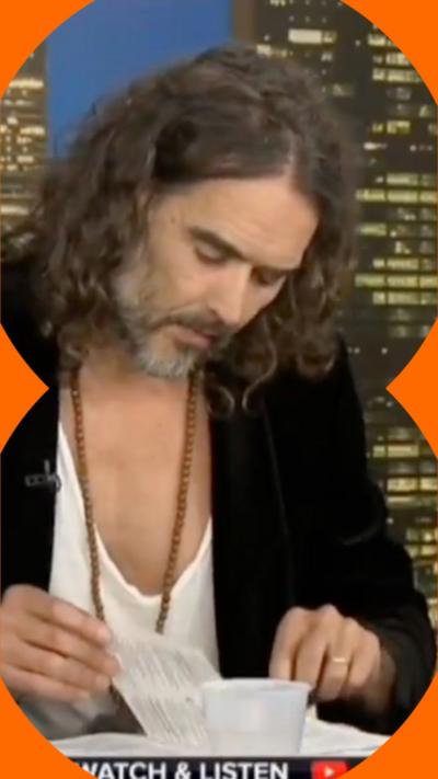 Russell Brand looks down at a Bible, flicking through the pages. He's wearing a black blazer and low-cut white t-shirt.