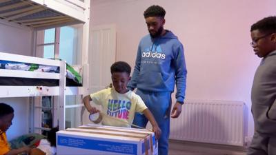 A child in a yellow shirt uses tape on a cardboard box while his father watches in a bedroom with bunk beds in the background.
