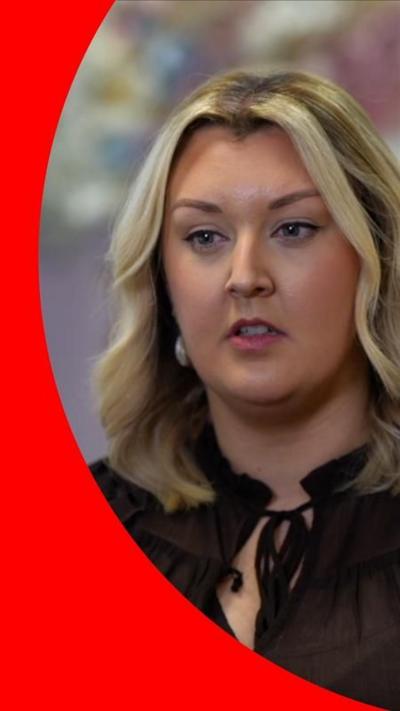 A woman with wavy brown hair with highlights looks off camera. She is wearing a black shirt tied at the neck