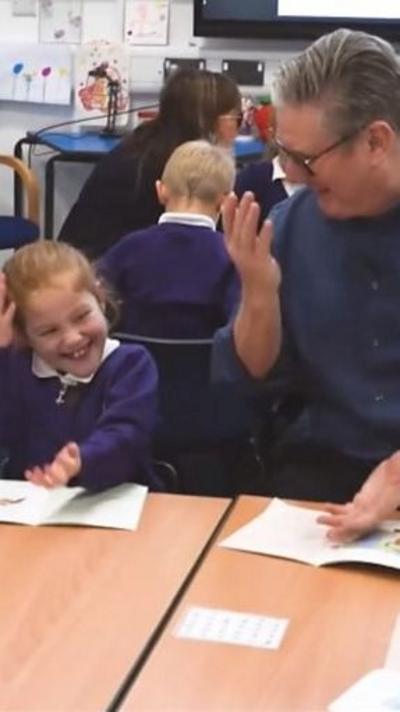 Starmer and a young pupil do the 6-7 hand gesture in a classroom