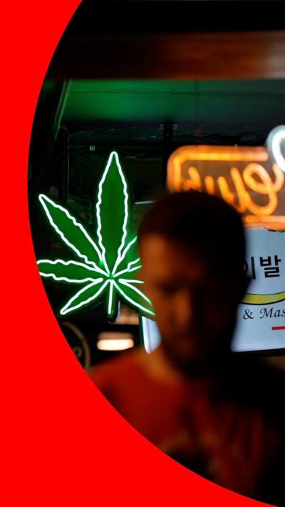 A man stands in front of a neon sign of a marijuana leaf