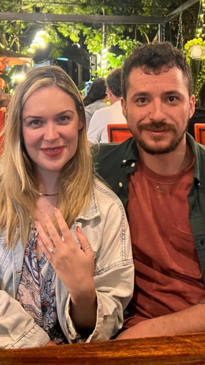 Man with brown hair, a dark green shirt and a faded orange t shirt sits next to a blonde woman with a small gold necklace and a faded Denim jacket. They are sitting in a crowded outdoor seating area of a restaurant or bar, there are green ornaments above their heads and leafy decorations. There are also bright orange chairs.