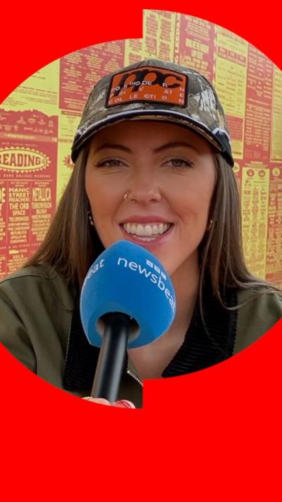Artist LYVIA has long brown hair and wears a baseball cap and a green jacket. She's holding a newsbeat mic