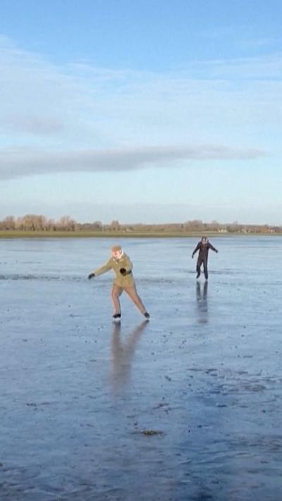 People skating on a frozen flood plane in Oxford - two figures can be seen in motion in the image - both in motion - the frozen lake and blue sky stretch out behind the two figures