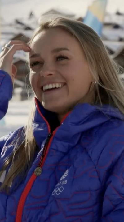 A woman with long blonde straight hair smiling as she brushes hair out of her face. She is wearing a blue jacket with the Team GB logo and Olympic rings on it.