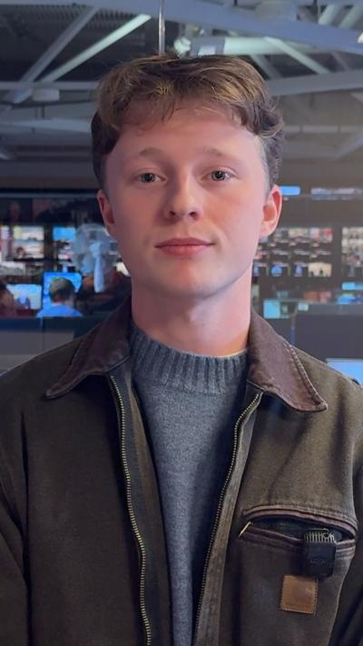 Jamie McColgan looking at the camera in the BBC NI newsroom.  He has short, fair hair and is wearing a brown jacket and a grey jumper.