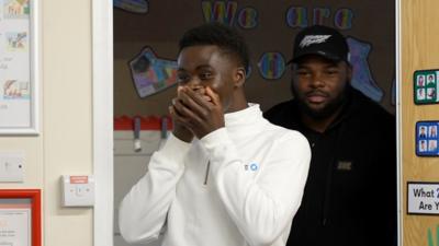 Bukayo Saka in a white zip-up shirt stands nervously, with hands covering his mouth, in a classroom setting.