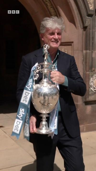 A man with grey hair in a blue suit with green shirt holding a silver football trophy with blue and white ribbons