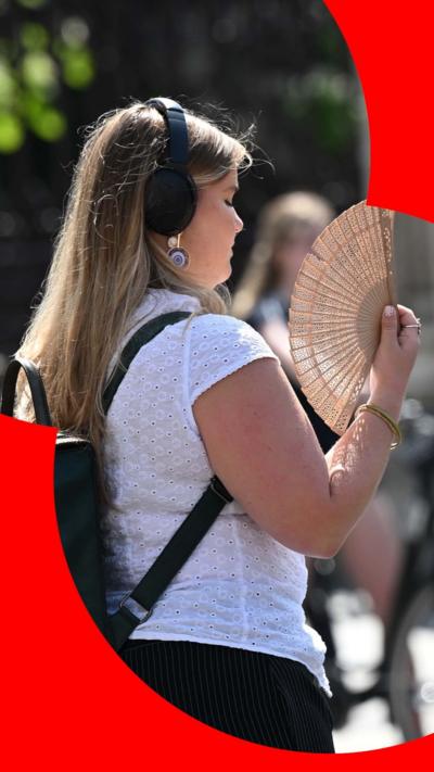Woman with long blonde hair, wearing headphones and white top uses a hand fan to cool herself down in the street.