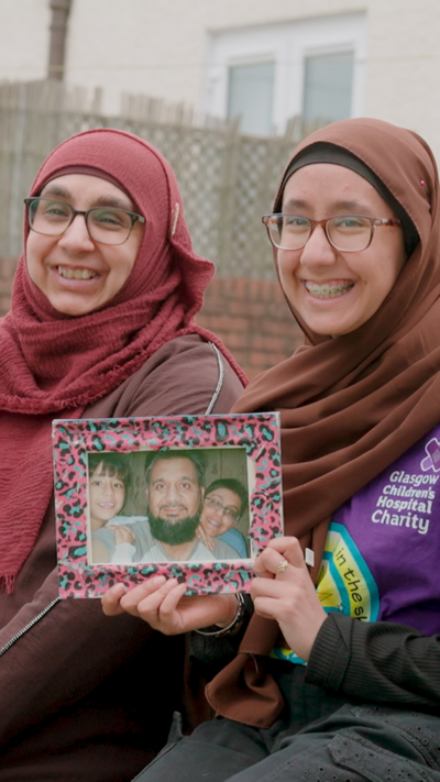 Two women wearing pink and brown hijabs, sit side-by-side holding a framed photo