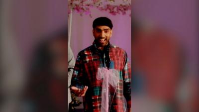 A man wearing a red tartan shirt, mid speech, in front of mic with a pink bow tied to it.