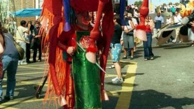 A photograph of a character in carnival outfit.  The whole outfit is not visible but the person wearing the outfit has a green dress or apron on and the costume appears to have a red horse headdress which engulfs the person below.  Behind members of the public can be seen standing and observing on the street.