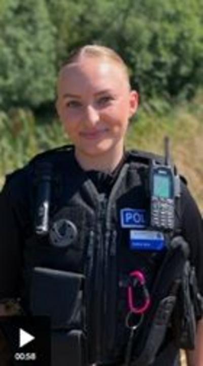A young woman dressed in a police uniform smiles at the camera. She has blonde hair tied back. Behind her are trees and grass
