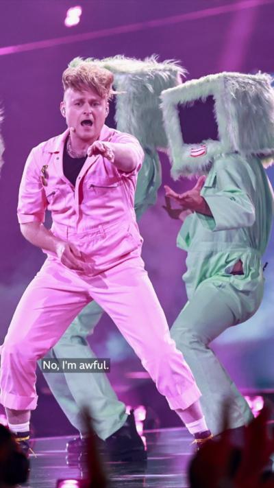 Man with messy blonde hair on stage wearing white top and trousers with dancers with giant TV heads behind him