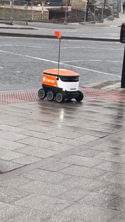 A Just Eat-branded orange and white delivery robot crossing a pedestrian crossing. It has six wheels and a pole reaching out from the top with an orange flag on the top. 