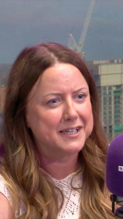 A woman with long brown curly hair wears a white lace top. She is smiling and sat in front of a purple microphone