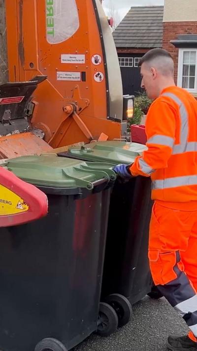 A young man with short brown hair with shaved sides loads two black bins into an orange refuse vehicle, about to be emptied. He is working in the street on a cloudy day. 