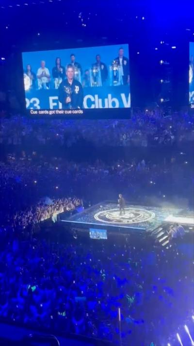 a picture of Noel Gallagher stood on a square stage in the Co-op Live arena, he is surrounded by thousands of people and the lighting is blue. He is holding a trophy and can be seen on stage