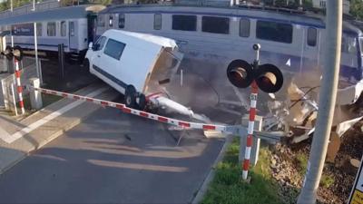 At a level crossing where red and white barriers are down, a blue and white train passes a damaged white van.