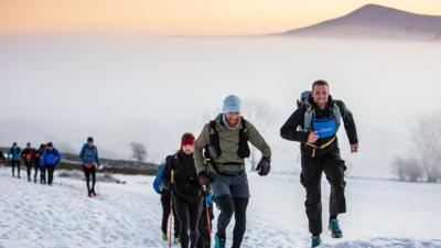 Racers run and trek through snow holding walking poles. They wear warm clothing. A mountain can be seen in background.