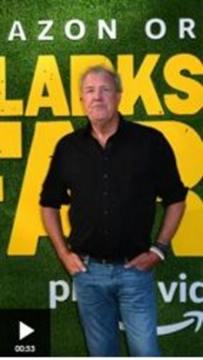 Jeremy Clarkson standing against a background of yellow text on a green background. He is wearing a black shirt and blue jeans, and has his hands in his pockets.