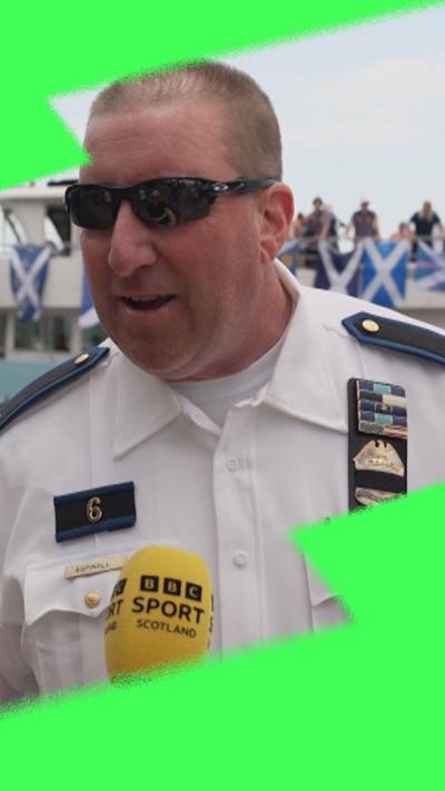 A police officer in his uniform and black sunglasses is speaking into a yellow 'BBC Sport Scotland' branded microphone.