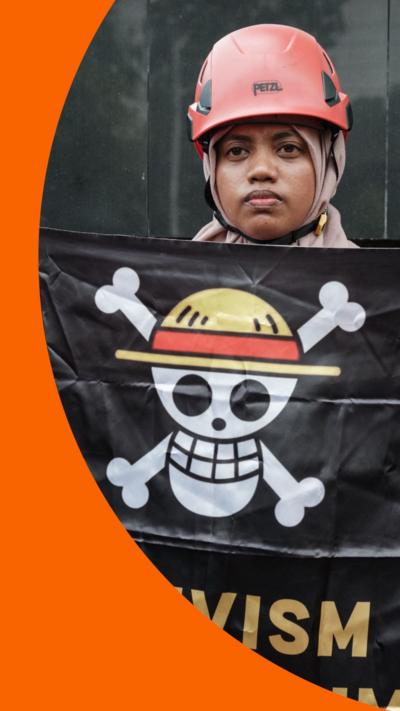 A protester in Indonesia holds a black flag with the One Piece pirate symbol - white skull and crossbones with a round yellow hat. She wears a safety helmet and headscarf, and looks stern
